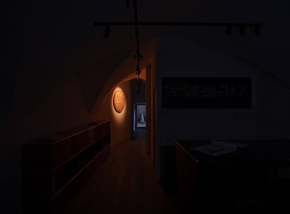 Dunkler Flur mit warmem Lichtakzent und Sicht auf einen beleuchteten Raum im Hintergrund.